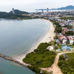 Vista aérea da costa de Santa Catarina, com faixa de praia, mar e área urbana à margem do litoral.