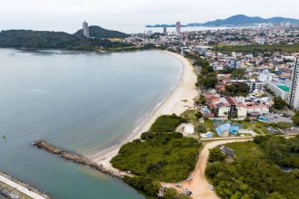 Vista aérea da costa de Santa Catarina, com faixa de praia, mar e área urbana à margem do litoral.