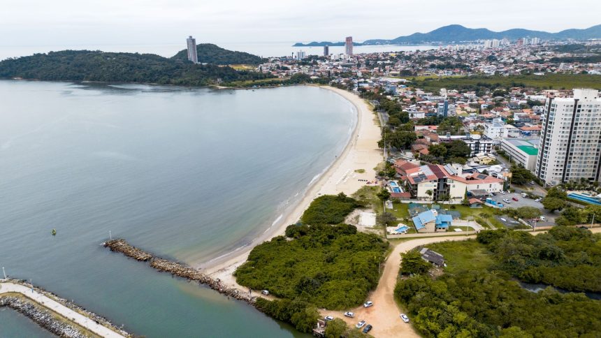 Vista aérea da costa de Santa Catarina, com faixa de praia, mar e área urbana à margem do litoral.