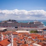 Navio de cruzeiro atracado diante de uma cidade litorânea, com telhados vermelhos e céu azul.