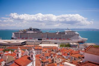 Navio de cruzeiro atracado diante de uma cidade litorânea, com telhados vermelhos e céu azul.
