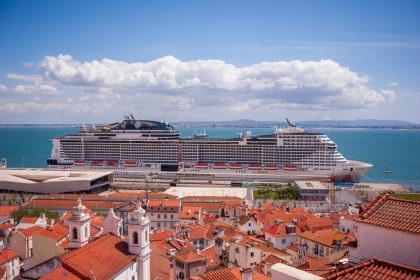 Navio de cruzeiro atracado diante de uma cidade litorânea, com telhados vermelhos e céu azul.