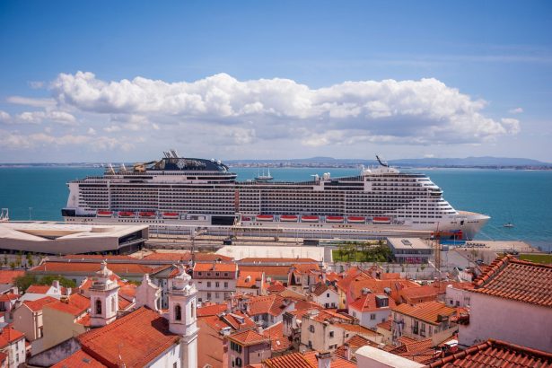 Navio de cruzeiro atracado diante de uma cidade litorânea, com telhados vermelhos e céu azul.