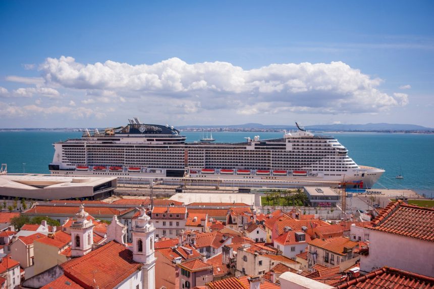 Navio de cruzeiro atracado diante de uma cidade litorânea, com telhados vermelhos e céu azul.