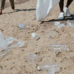 Resíduos plásticos espalhados na areia enquanto pessoas fazem a limpeza de uma praia.