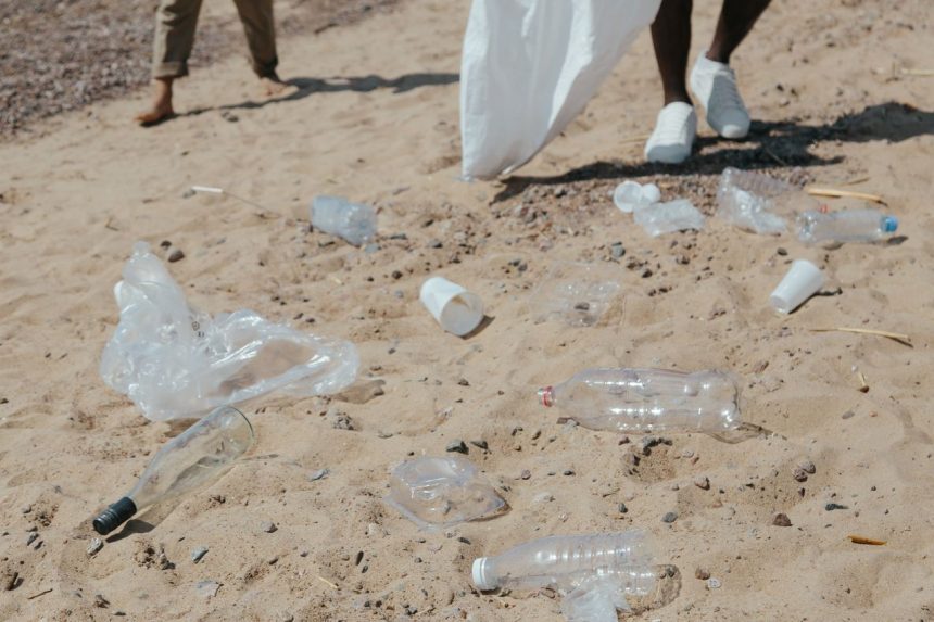 Resíduos plásticos espalhados na areia enquanto pessoas fazem a limpeza de uma praia.