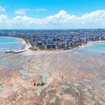 Vista aérea da orla de Maceió, com faixa de praia, águas turquesa e área urbana ao longo da costa.