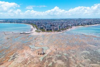 Vista aérea da orla de Maceió, com faixa de praia, águas turquesa e área urbana ao longo da costa.