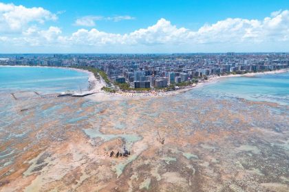 Vista aérea da orla de Maceió, com faixa de praia, águas turquesa e área urbana ao longo da costa.