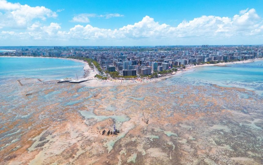 Vista aérea da orla de Maceió, com faixa de praia, águas turquesa e área urbana ao longo da costa.