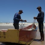 Dois pescadores preparam redes junto a um barco colorido na praia de Itaúnas, no Espírito Santo.