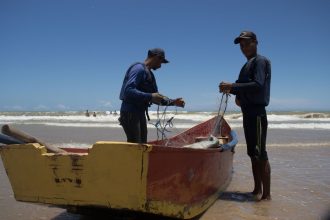 Dois pescadores preparam redes junto a um barco colorido na praia de Itaúnas, no Espírito Santo.