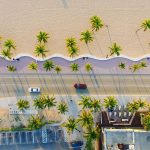 Vista aérea de uma área litorânea urbanizada, com praia, coqueiros, edificações, estrada e veículos junto à faixa de areia.