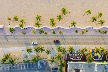 Vista aérea de uma área litorânea urbanizada, com praia, coqueiros, edificações, estrada e veículos junto à faixa de areia.