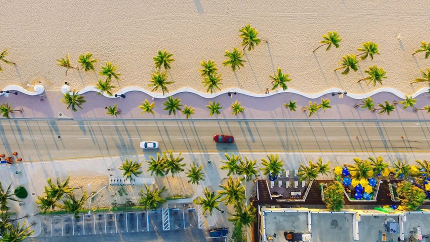 Vista aérea de uma área litorânea urbanizada, com praia, coqueiros, edificações, estrada e veículos junto à faixa de areia.